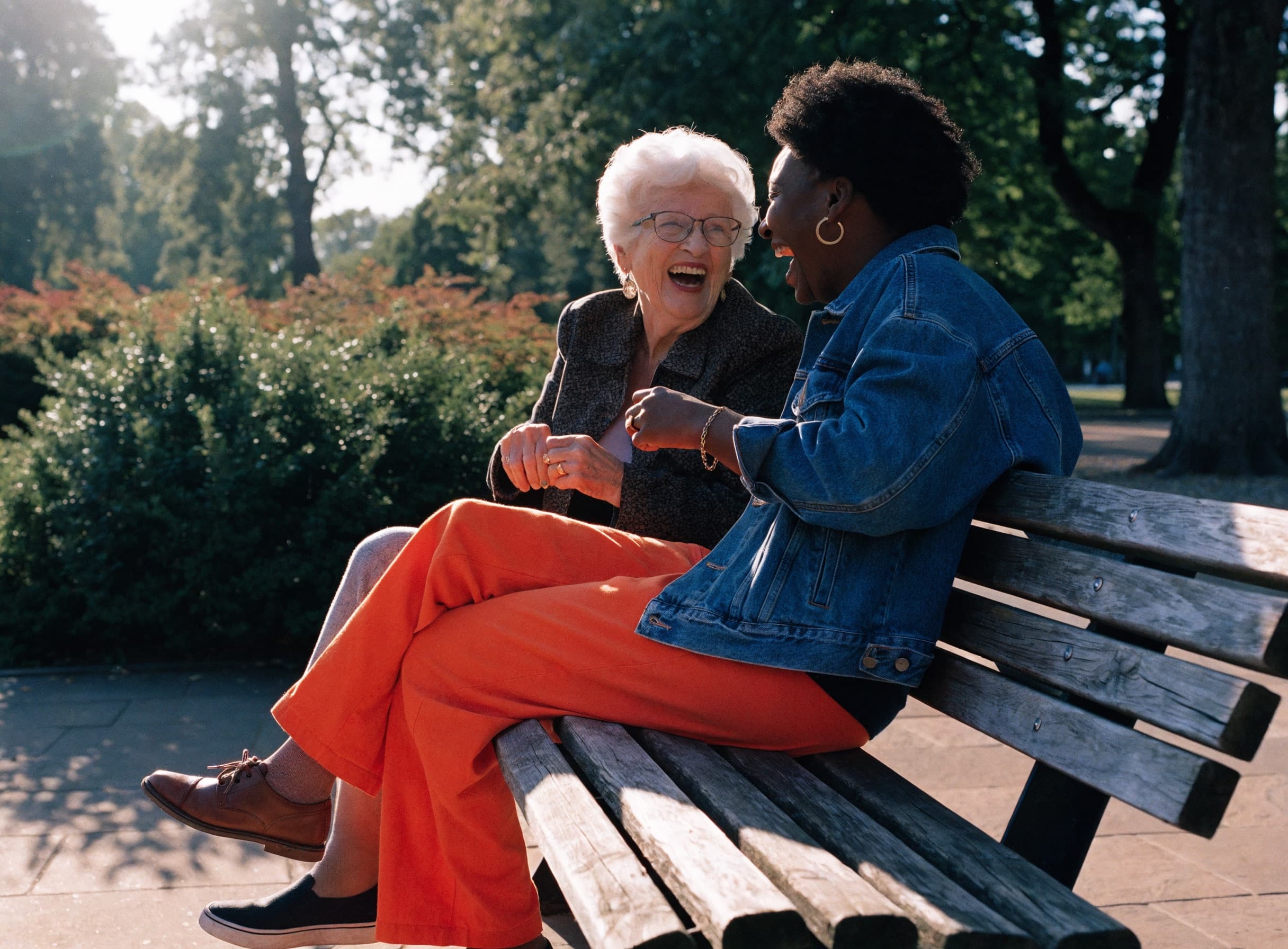An elderly woman and a companion carer laughing together on a park bench in the afternoon sun